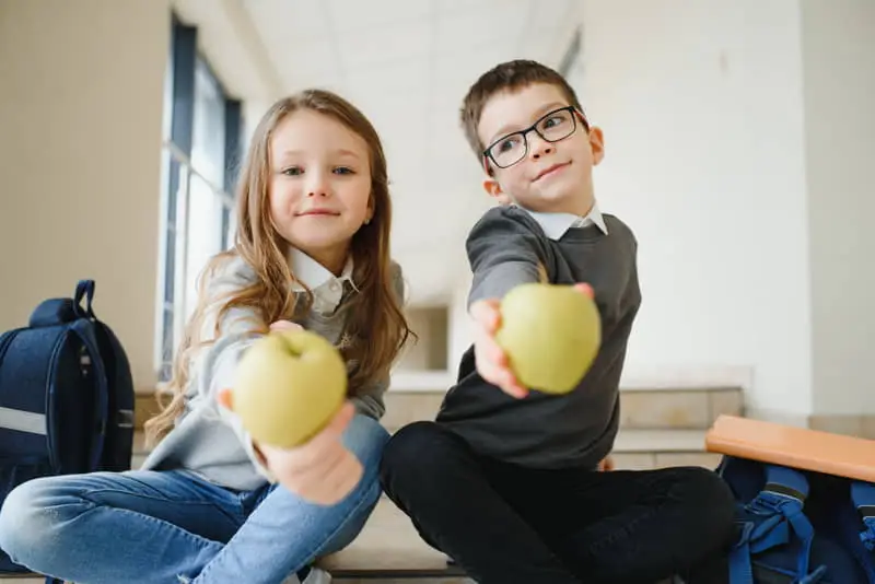 Dos niños sentados en escaleras interiores, sosteniendo manzanas verdes hacia la cámara, con mochilas escolares a su lado.