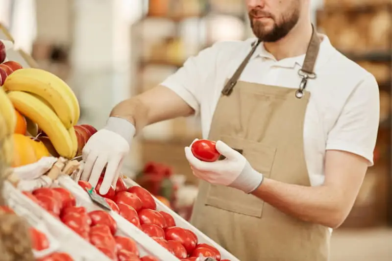 Hombre con delantal y guantes seleccionando tomates frescos en un mercado o tienda de frutas y verduras.