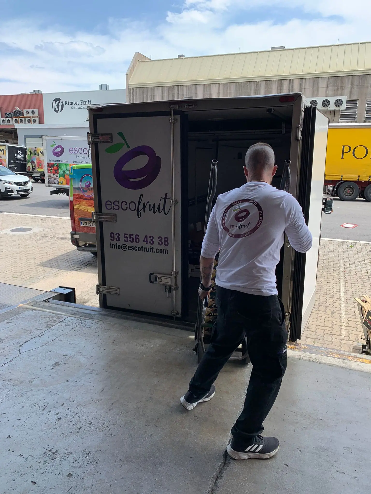 Hombre de espaldas con camiseta blanca y pantalones oscuros cargando cajas hacia una furgoneta de Escofruit abierta en un área de carga.